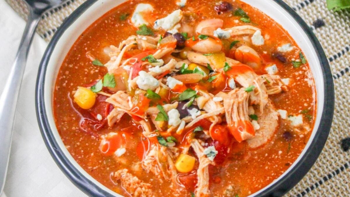 A bowl of chicken tortilla soup topped with crumbled cheese, chopped cilantro, corn, beans, and diced tomatoes—perfect for fans of zesty recipes.
