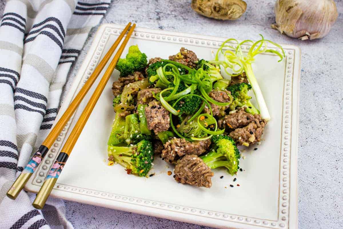 A square white plate with beef and broccoli stir-fry, garnished with scallions. Chopsticks rest on the side; a striped napkin and ginger root are visible in the background—a flavorful favorite among beef skillet meals.