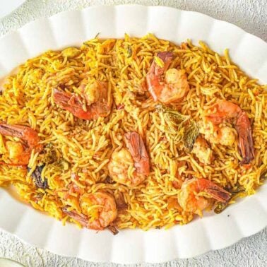 A white oval plate filled with shrimp biryani, featuring yellow rice, visible shrimp, and herbs—an epic rice bowl placed on a light textured surface.