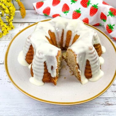 A bundt cake with white icing is on a plate, with a slice removed. Perfect for busy days desserts, it sits beside a floral napkin and yellow flowers on a white wooden surface.
