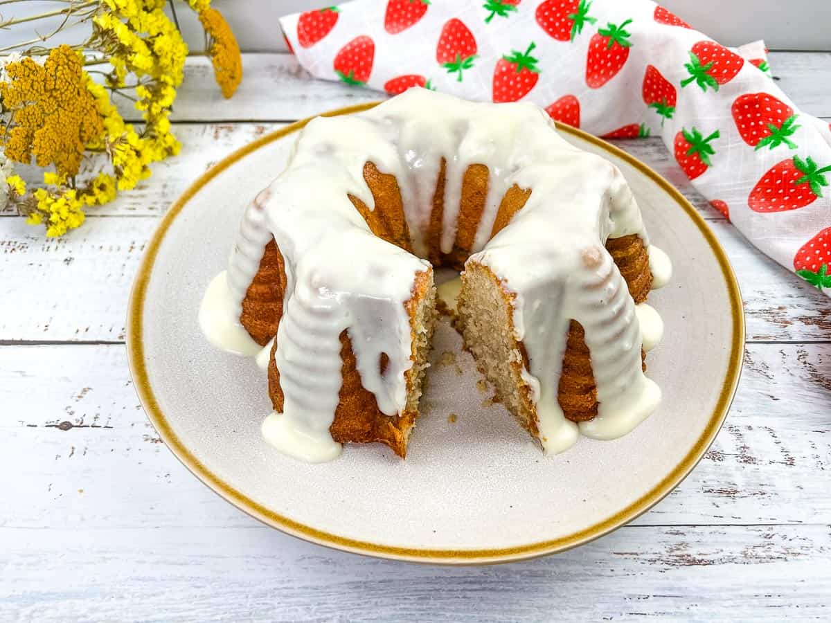 A bundt cake with white icing is on a plate, with a slice removed. Perfect for busy days desserts, it sits beside a floral napkin and yellow flowers on a white wooden surface.