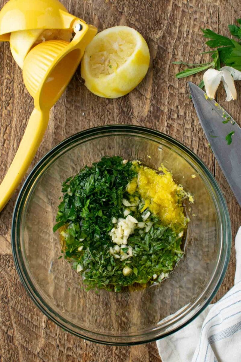 A glass bowl with chopped herbs, minced garlic, lemon zest, and olive oil on a wooden surface, next to a knife, a cut lemon, and a yellow citrus squeezer.