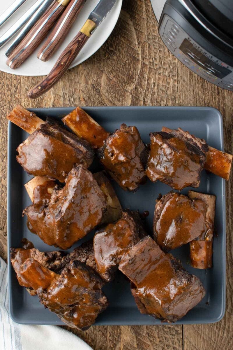A gray plate filled with braised beef short ribs covered in brown sauce sits on a wooden table next to an Instant Pot and a plate with utensils.