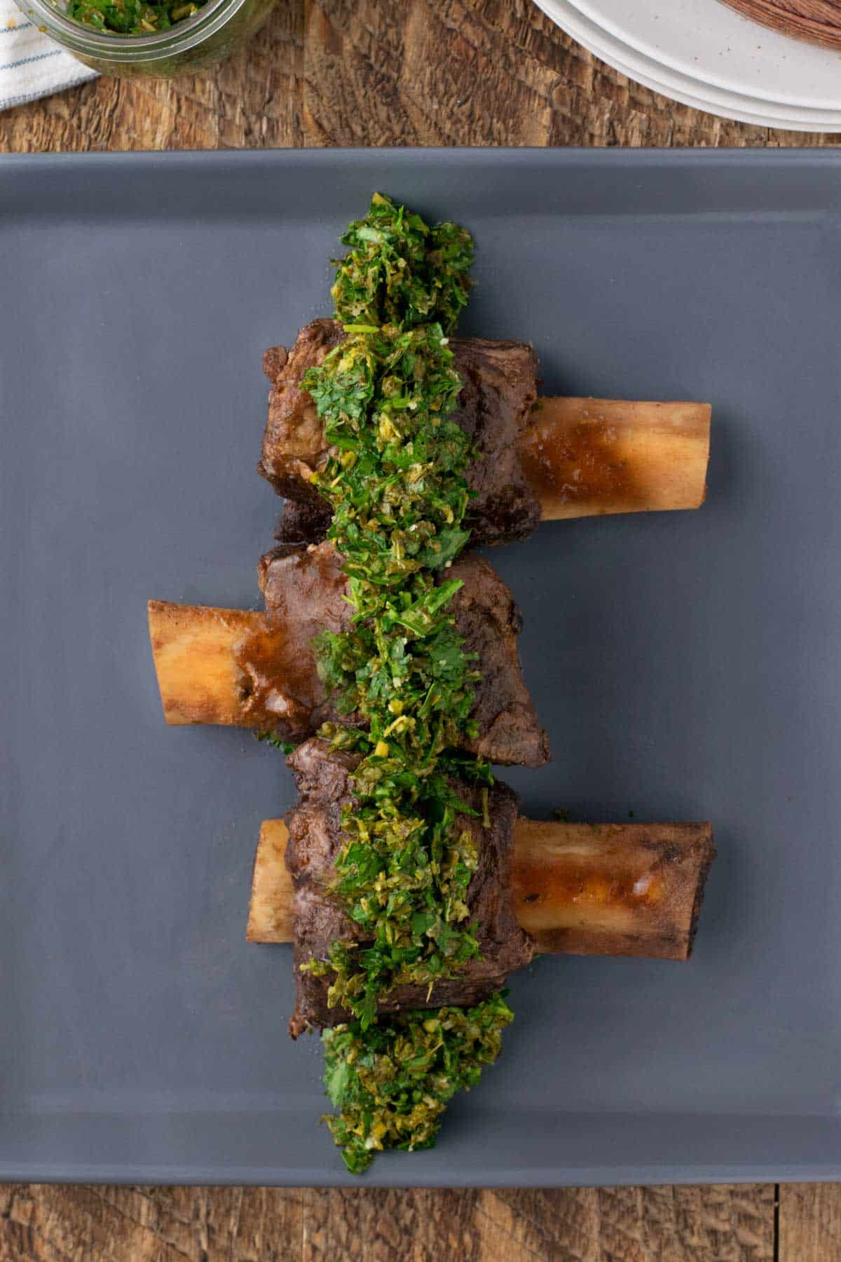 Three cooked beef short ribs arranged in a row on a gray plate, topped with a green herb chimichurri sauce.