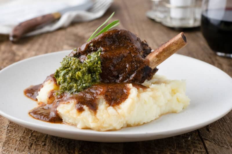 A plate of mashed potatoes topped with instant pot beef short ribs in brown sauce, garnished with green herb pesto, served on a wooden table.