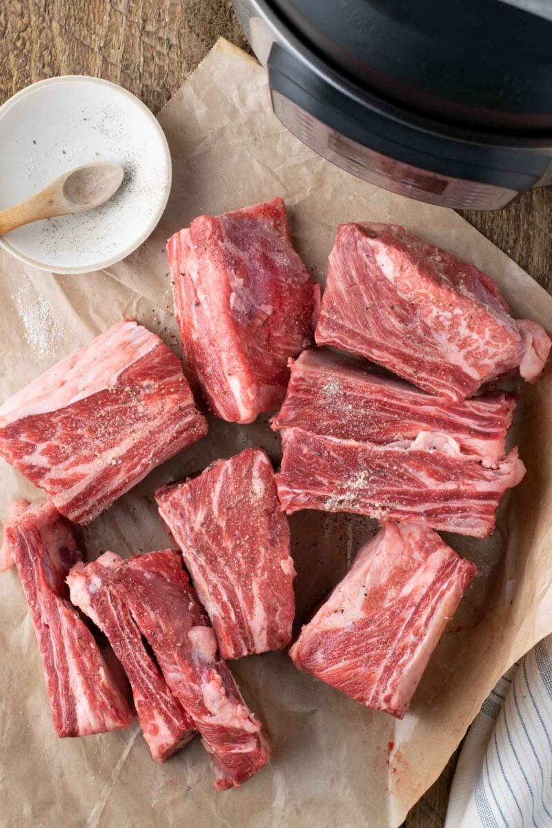 Raw beef short ribs are arranged on a sheet of parchment paper next to a small dish of salt and a kitchen appliance.