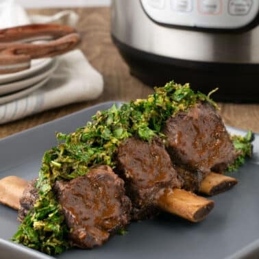 Three instant pot beef short ribs topped with chopped herbs are served on a gray plate, with an Instant Pot and a stack of plates in the background.
