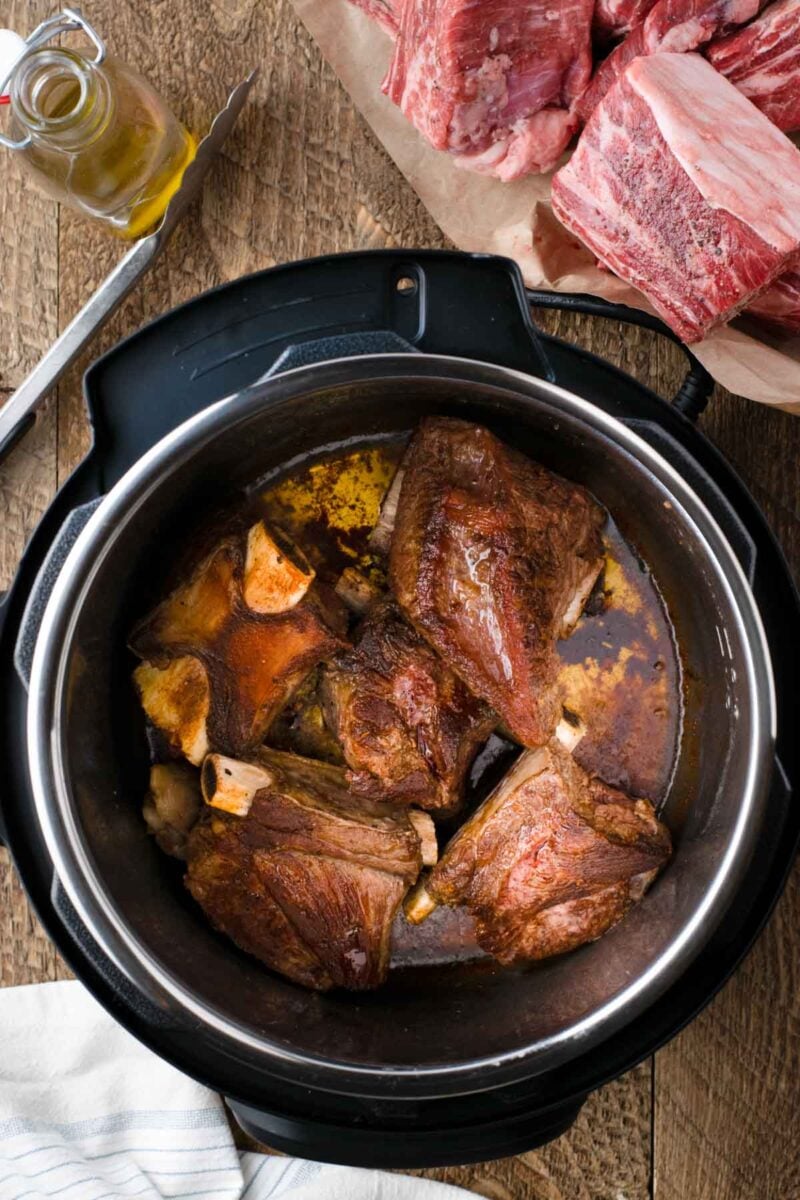 Four browned beef short ribs are searing in an Instant Pot, with raw ribs and a jar of oil on the wooden countertop nearby.