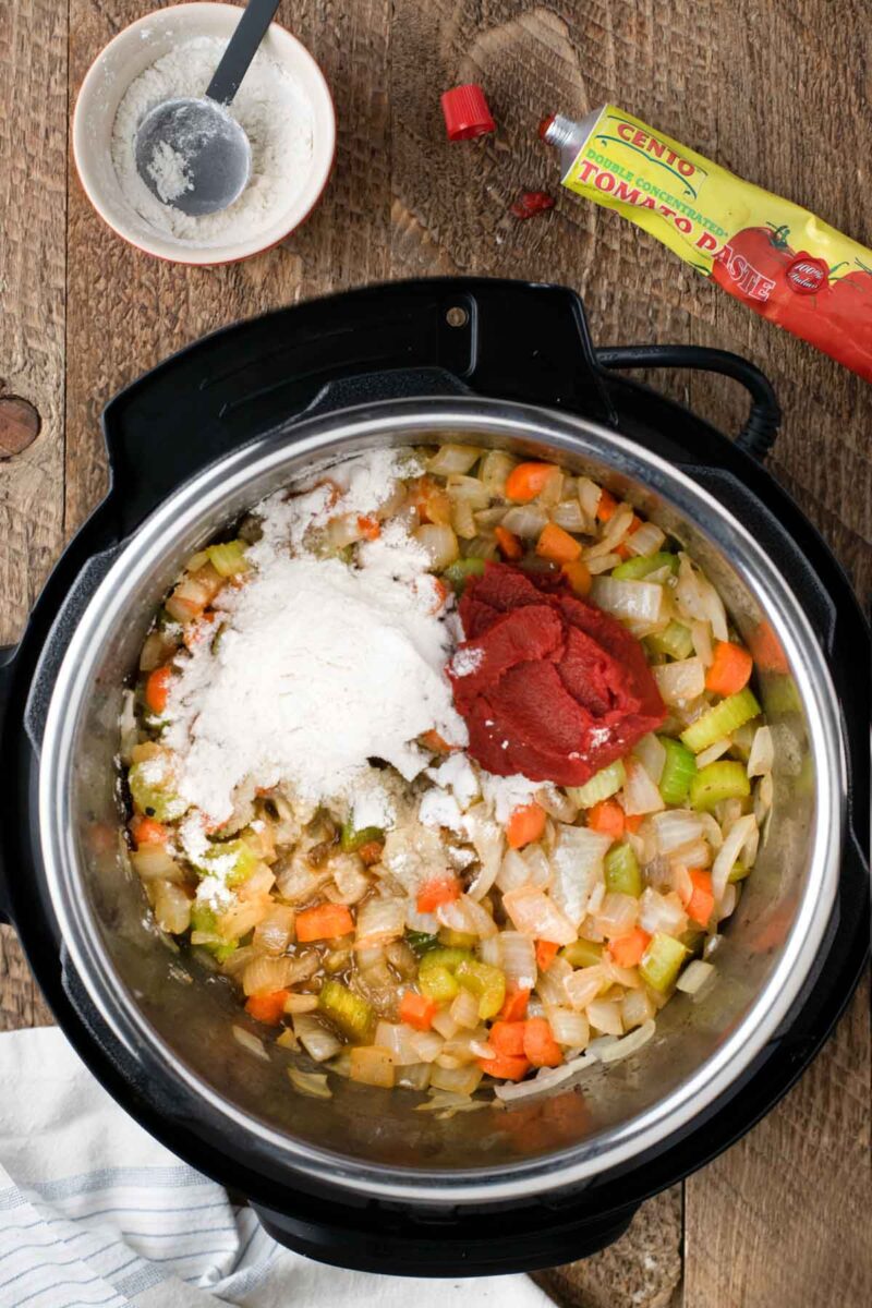 Overhead view of an Instant Pot with sautéed onions, carrots, and celery, topped with a scoop of flour and a dollop of tomato paste; ingredients and tools nearby.