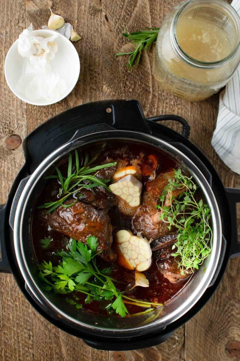 An overhead view of an Instant Pot containing beef, fresh herbs, and halved garlic bulbs in broth, with a jar of stock and a bowl of garlic cloves on a wooden surface nearby.