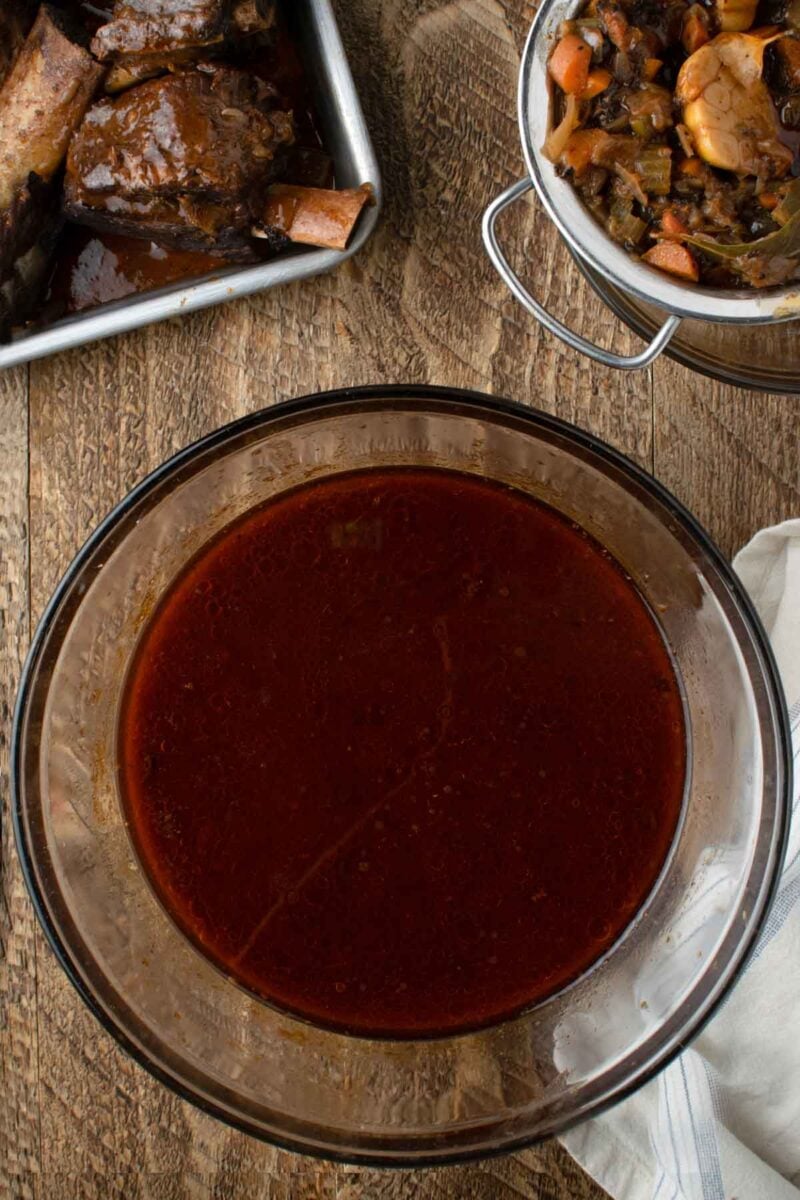 A glass bowl filled with brown beef au jus sits on a wooden table, with a tray of cooked short ribs and a bowl of vegetables nearby.