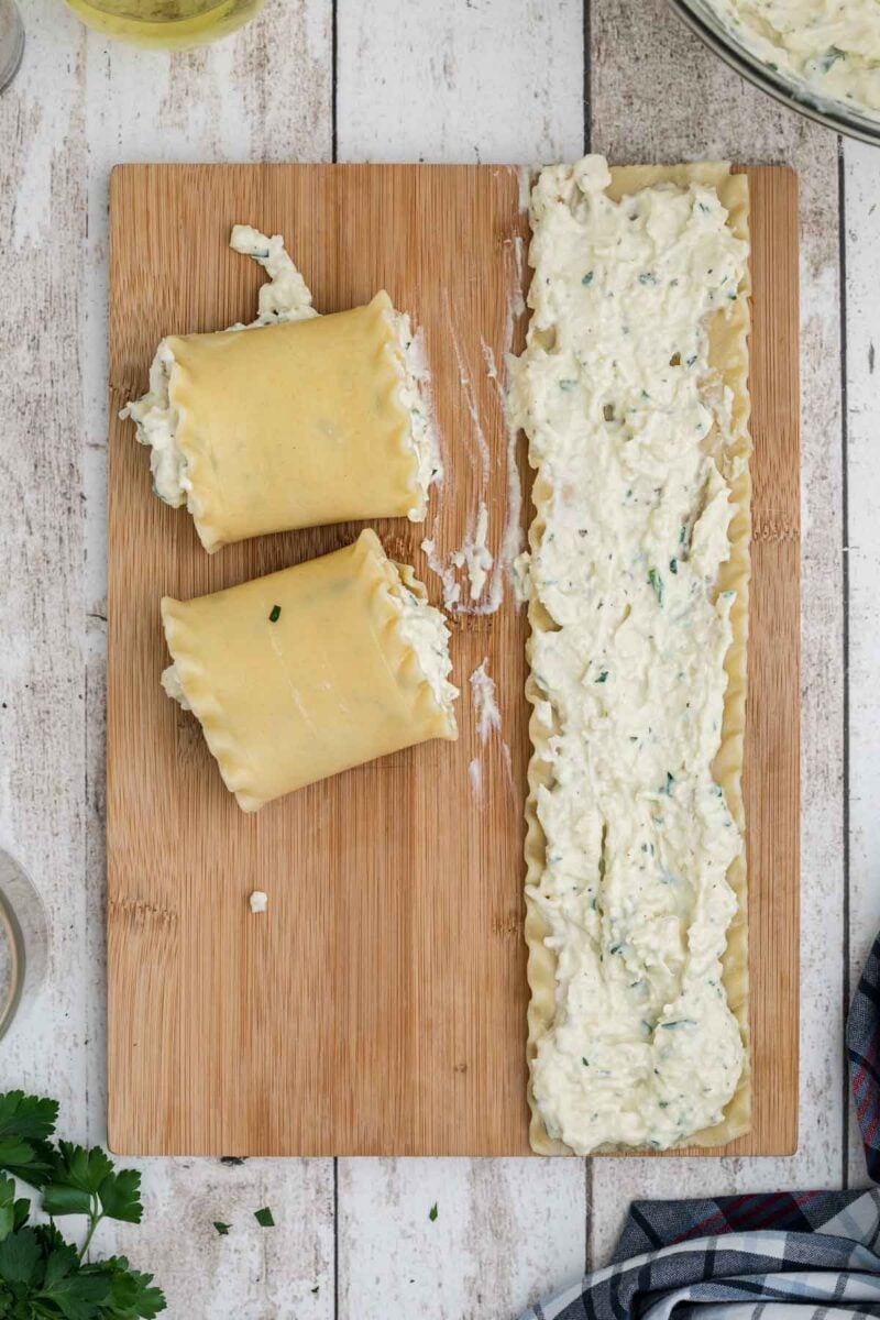 Two rolled lasagna noodles and one unrolled noodle with cheese filling on a wooden cutting board, viewed from above.