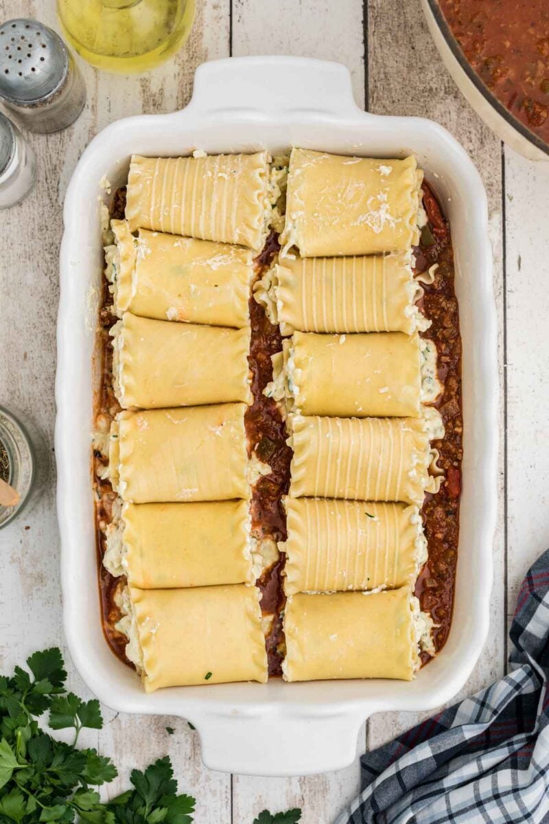 A white baking dish filled with rolled lasagna noodles placed on top of a layer of meat sauce, ready to be baked.