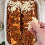 A hand sprinkles shredded cheese over a partially assembled lasagna in a white baking dish on a wooden surface.