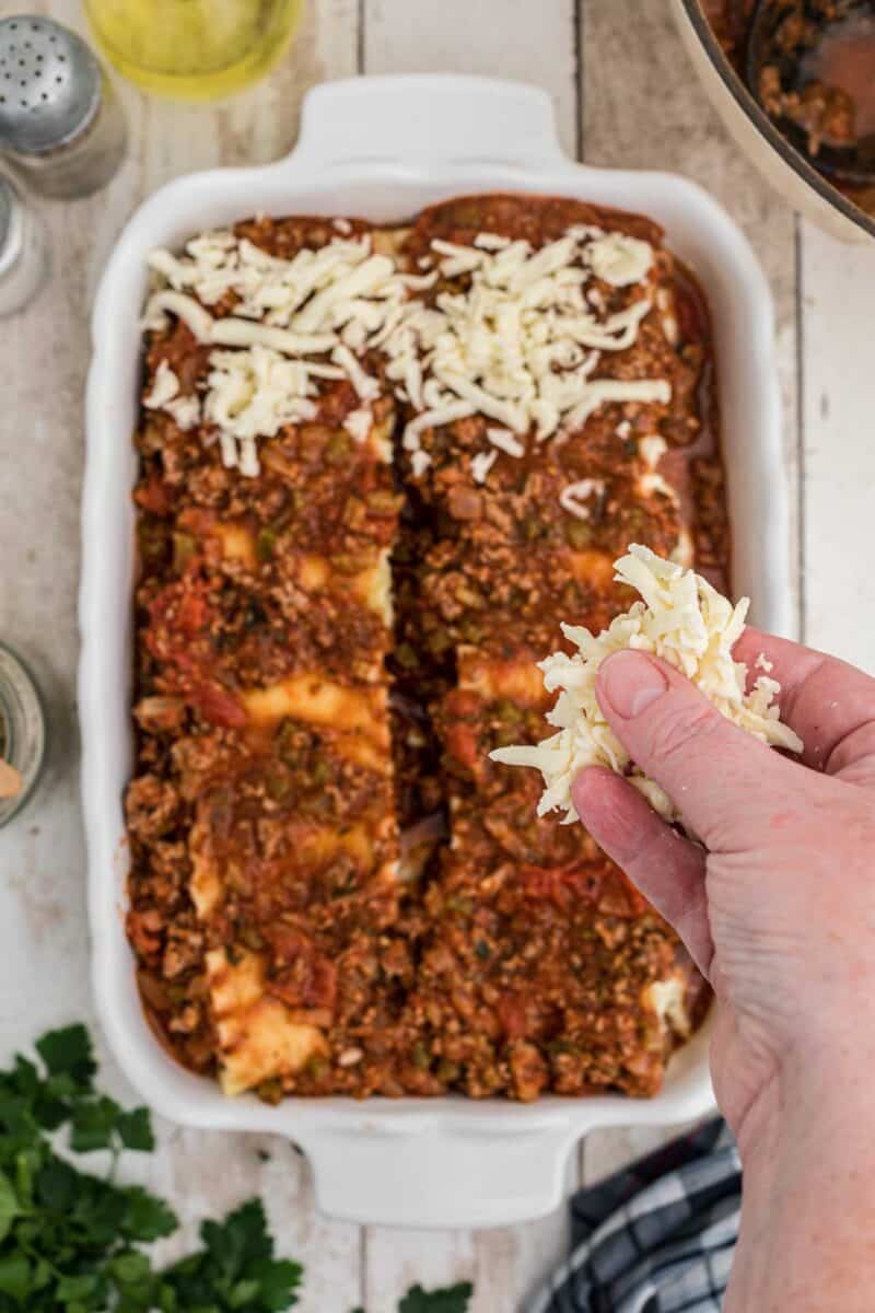 A hand sprinkles shredded cheese over a partially assembled lasagna in a white baking dish on a wooden surface.