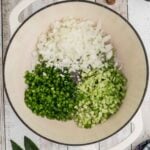 A large pot with chopped onions, green bell peppers, and celery arranged in sections, ready to be cooked, on a wooden surface with various ingredients nearby.