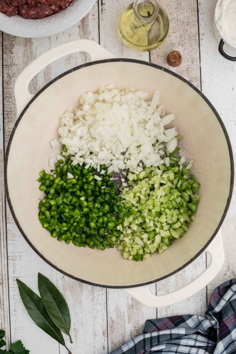 A large pot with chopped onions, green bell peppers, and celery arranged in sections, ready to be cooked, on a wooden surface with various ingredients nearby.