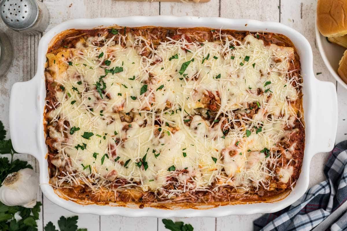 A baked lasagna rollup in a white dish topped with melted cheese and sprinkled with chopped parsley, shown on a white wooden surface.