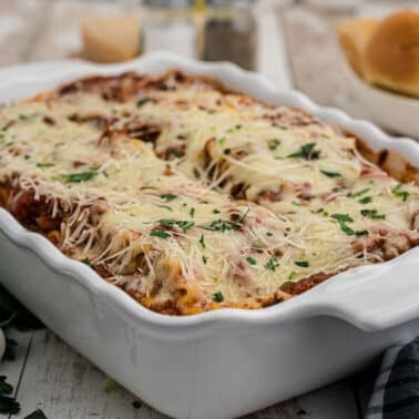 A baked lasagna rollups topped with melted cheese and parsley in a white dish, with bread rolls and fresh garlic nearby on a wooden table.