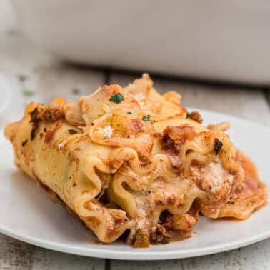 A piece of lasagna roll-up filled with ricotta and meat sauce sits on a white plate, garnished with herbs on a wooden table.