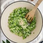 Diced onions, celery, and green bell peppers are sautéing in a white pot with minced garlic added on top; a wooden spoon rests to the side.