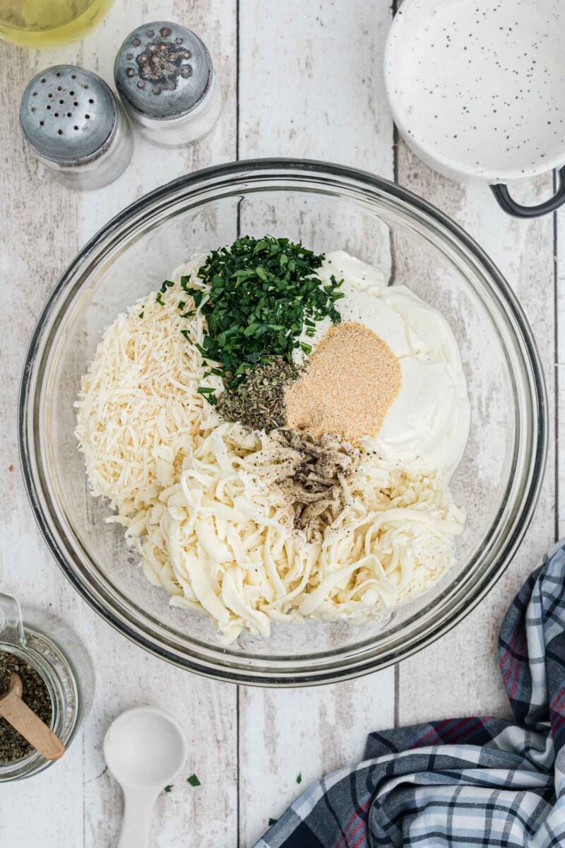 A glass bowl filled with shredded cheese, chopped parsley, dried herbs, garlic powder, and ricotta cheese on a white wooden surface with nearby seasonings and utensils.