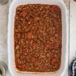 A white baking dish filled with cooked ground meat and tomato sauce on a wooden surface, surrounded by kitchen items.