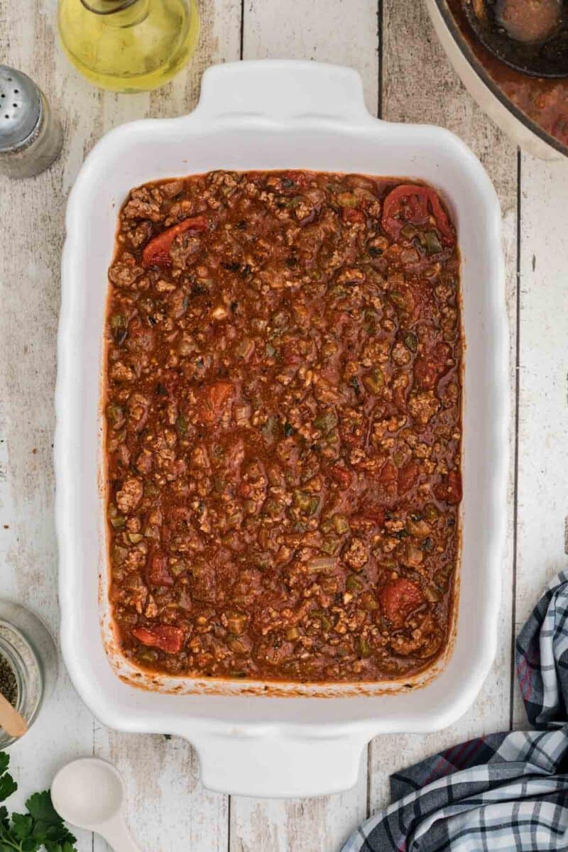 A white baking dish filled with cooked ground meat and tomato sauce on a wooden surface, surrounded by kitchen items.