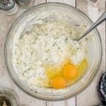 A glass bowl containing ricotta mixture and two raw eggs on a wooden surface, surrounded by seasonings, herb, a dish with eggshells, and a checked cloth.