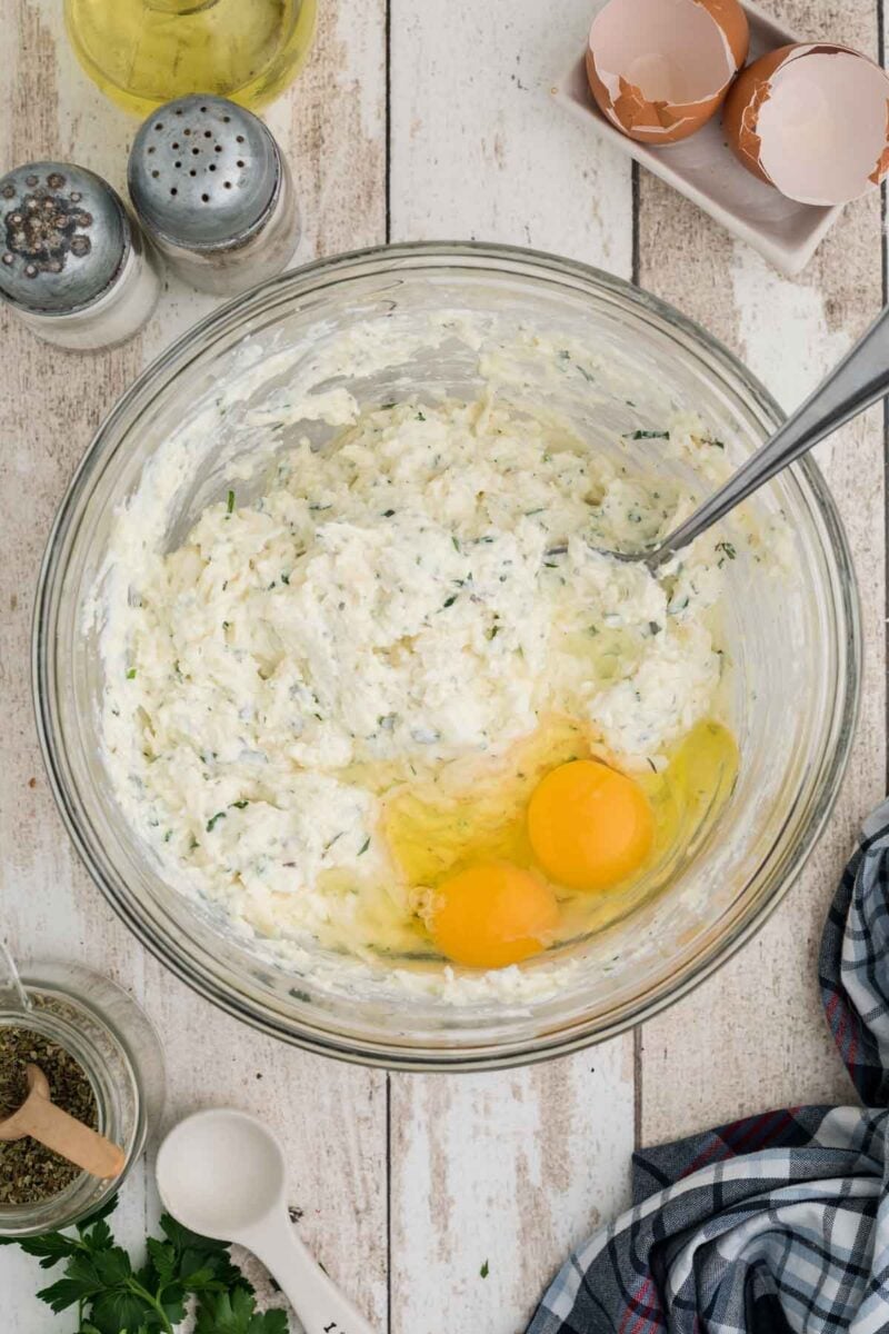A glass bowl containing ricotta mixture and two raw eggs on a wooden surface, surrounded by seasonings, herb, a dish with eggshells, and a checked cloth.