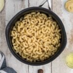 A black saucepan filled with cooked elbow macaroni sits on a white wooden surface surrounded by various small bowls and utensils.