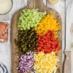 Chopped celery, yellow bell pepper, red bell pepper, pickles, red onion, and hard-boiled eggs arranged on a wooden cutting board with condiments nearby.