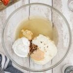 A glass bowl with sour cream, mayonnaise, mustard, spices, and vinegar, next to chopped bell peppers on a wooden surface with measuring utensils nearby.