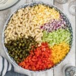 A large bowl containing separate sections of cooked macaroni, chopped hard-boiled egg, red onion, celery, yellow bell pepper, red bell pepper, and pickles.