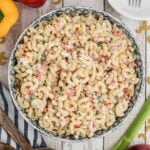 A bowl of Southern macaroni salad with diced vegetables sits on a wooden table, surrounded by celery, bell peppers, a red onion, and serving utensils.