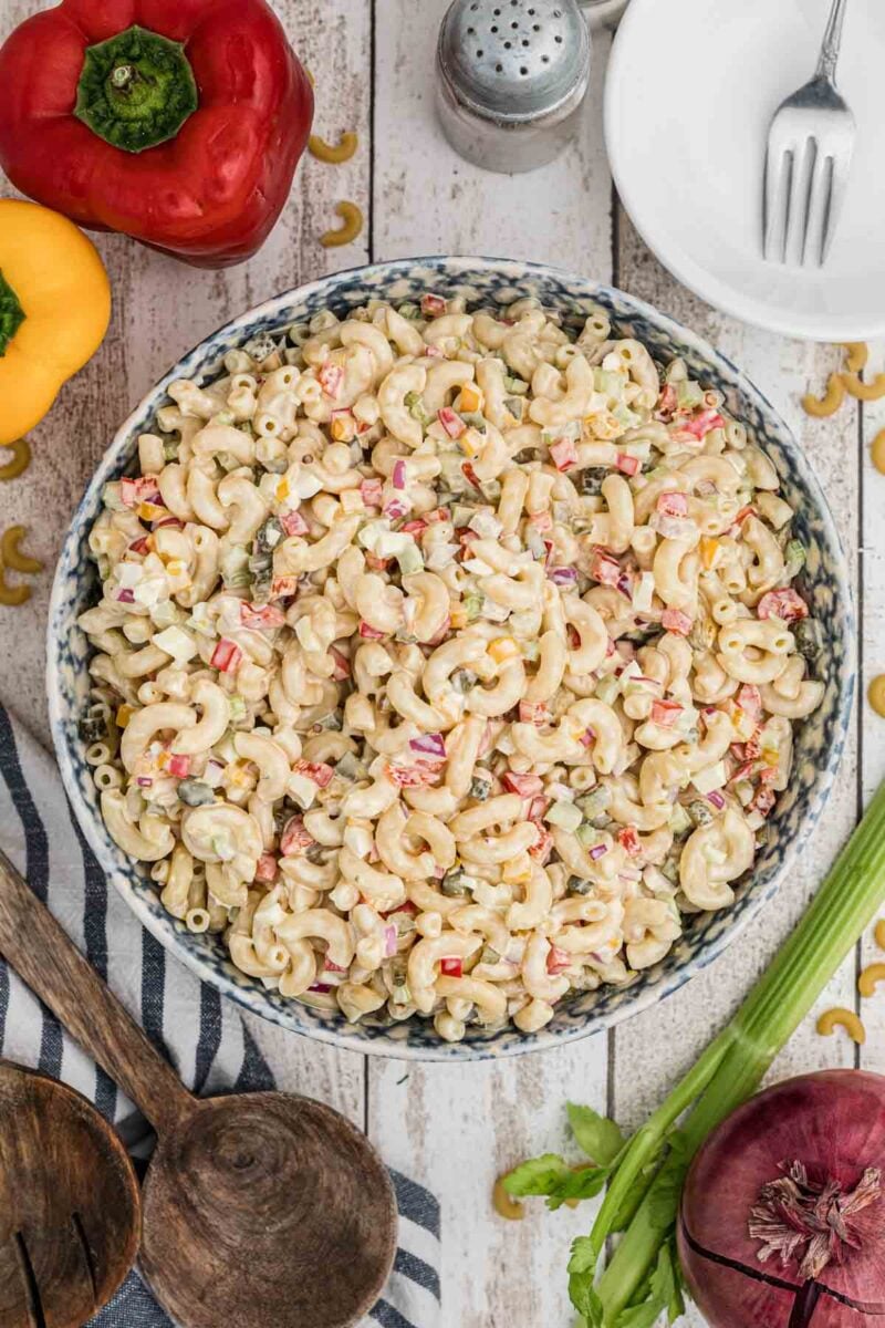 A bowl of Southern macaroni salad with diced vegetables sits on a wooden table, surrounded by celery, bell peppers, a red onion, and serving utensils.