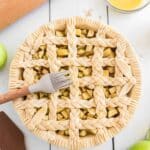 Unbaked apple pie with a braided lattice crust on a white table, surrounded by a rolling pin, egg wash, spatula, and green apples.
