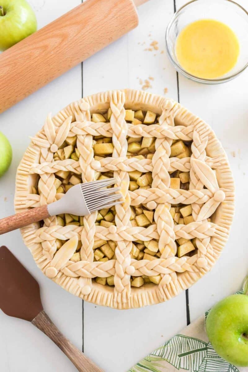 Unbaked apple pie with a braided lattice crust on a white table, surrounded by a rolling pin, egg wash, spatula, and green apples.