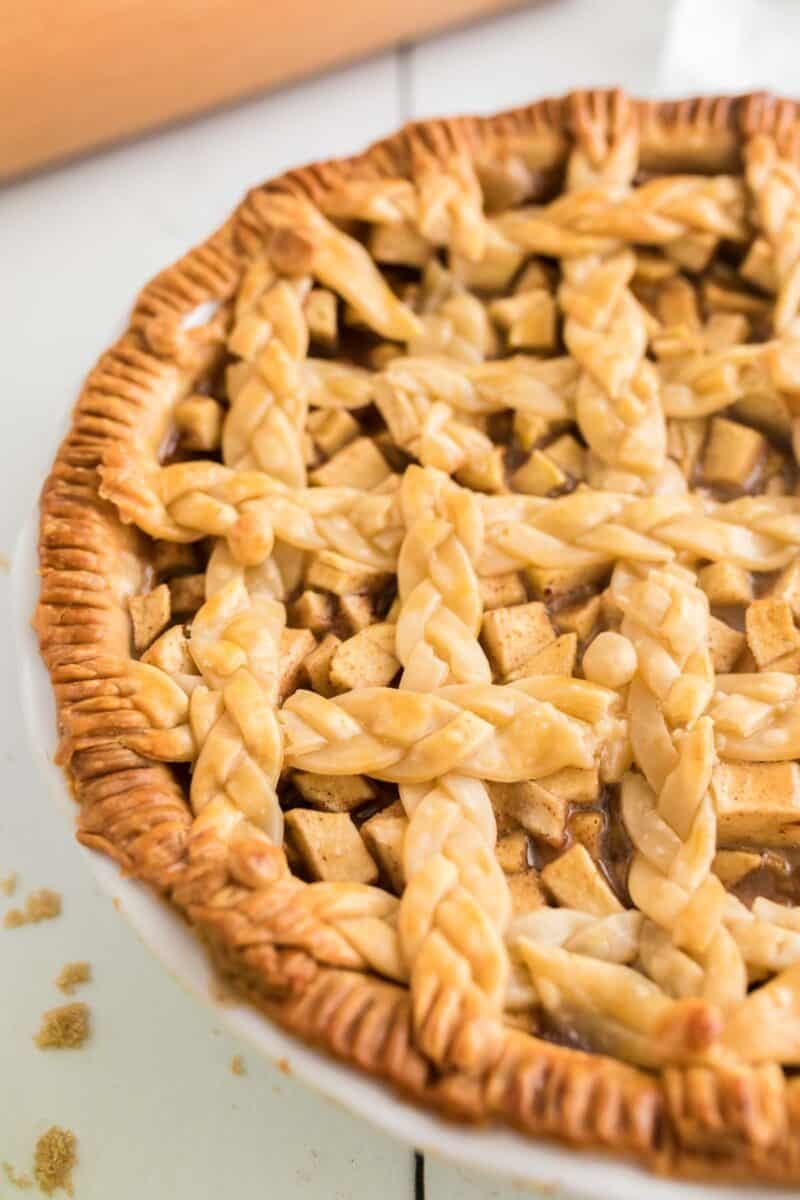 A close-up of a baked salted caramel apple pie with a golden brown, braided lattice crust in a white pie dish.