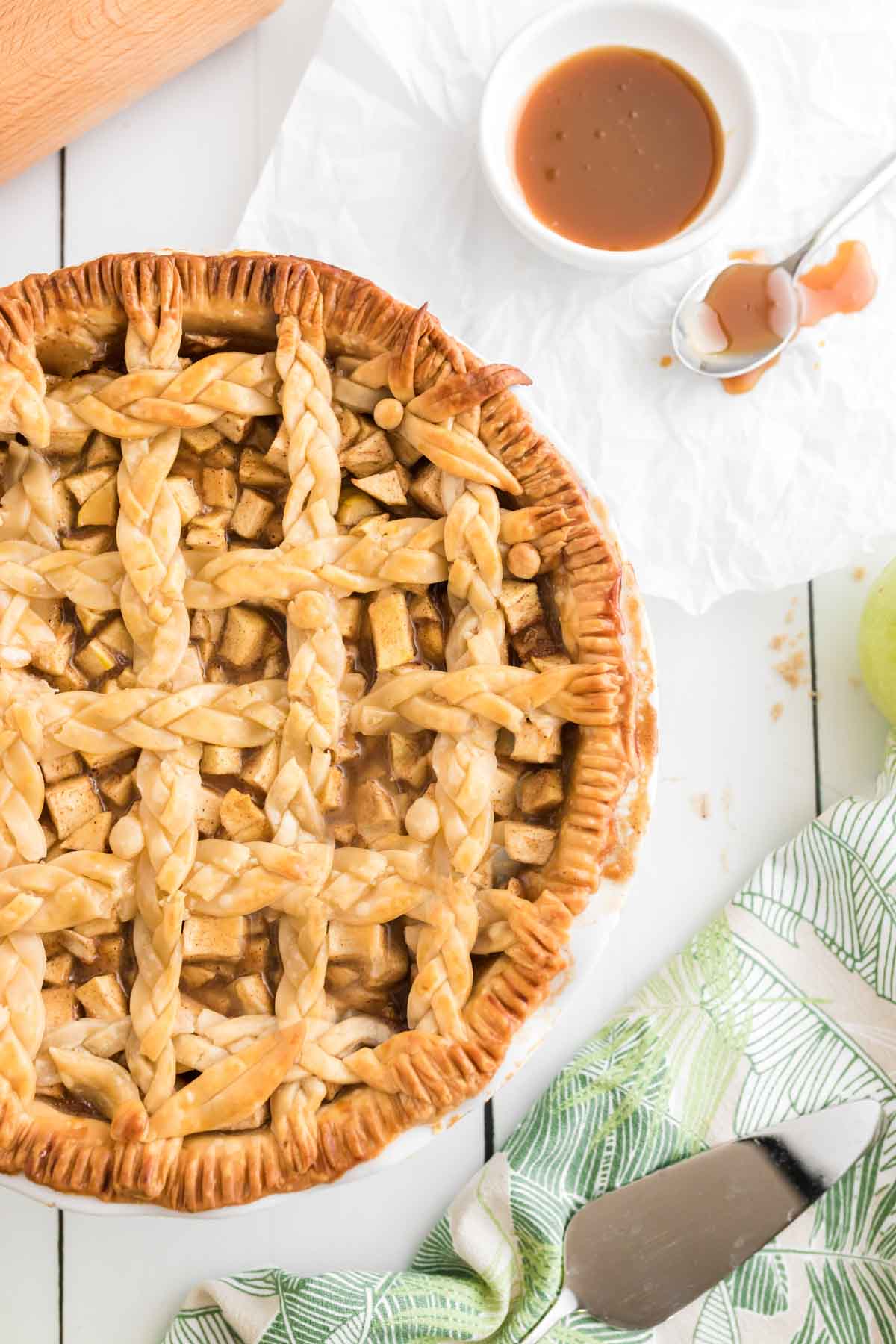 A lattice-topped salted caramel apple pie sits on a table next to a small bowl of caramel sauce, a spoon, a pie server, and a green-patterned cloth.