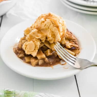 A slice of salted caramel apple pie topped with a scoop of vanilla ice cream sits on a white plate with a fork on the side.