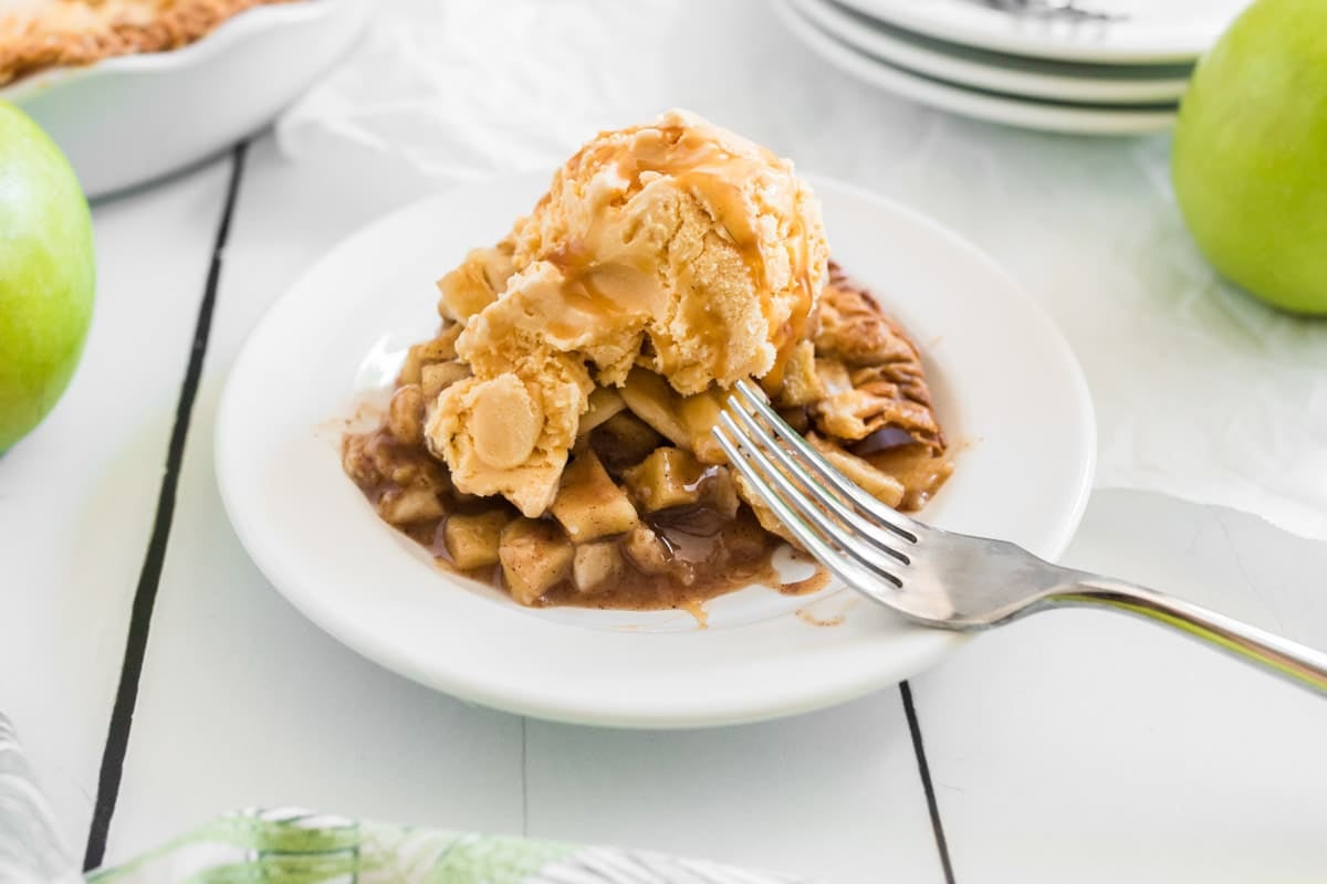 A slice of salted caramel apple pie topped with a scoop of vanilla ice cream sits on a white plate with a fork on the side.