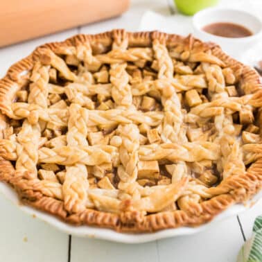 A baked salted caramel apple pie with a decorative lattice crust sits on a white plate, surrounded by baking utensils and ingredients on a white wooden table.