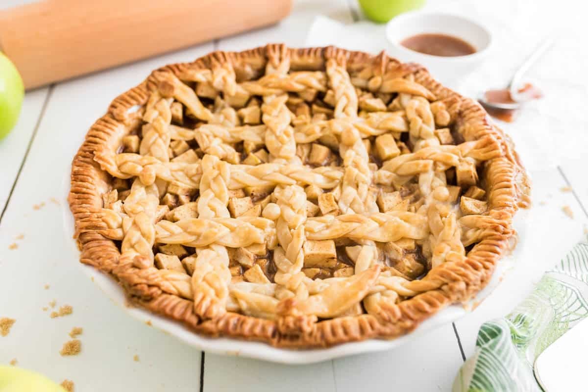 A baked salted caramel apple pie with a decorative lattice crust sits on a white plate, surrounded by baking utensils and ingredients on a white wooden table.