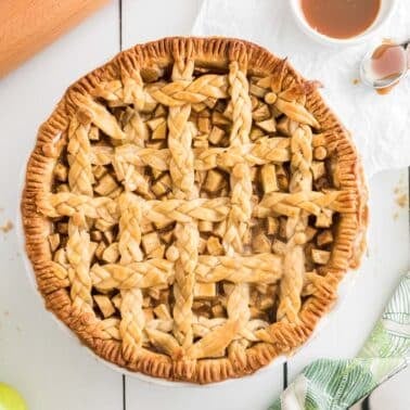 A baked salted caramel apple pie with a lattice crust sits on a white surface, surrounded by green apples, a rolling pin, a napkin, and a spoon with caramel sauce.