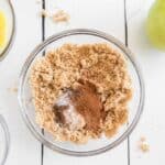 A glass bowl filled with brown sugar, cinnamon, and salt sits on a white surface, surrounded by other baking ingredients, including a green apple and a measuring cup.