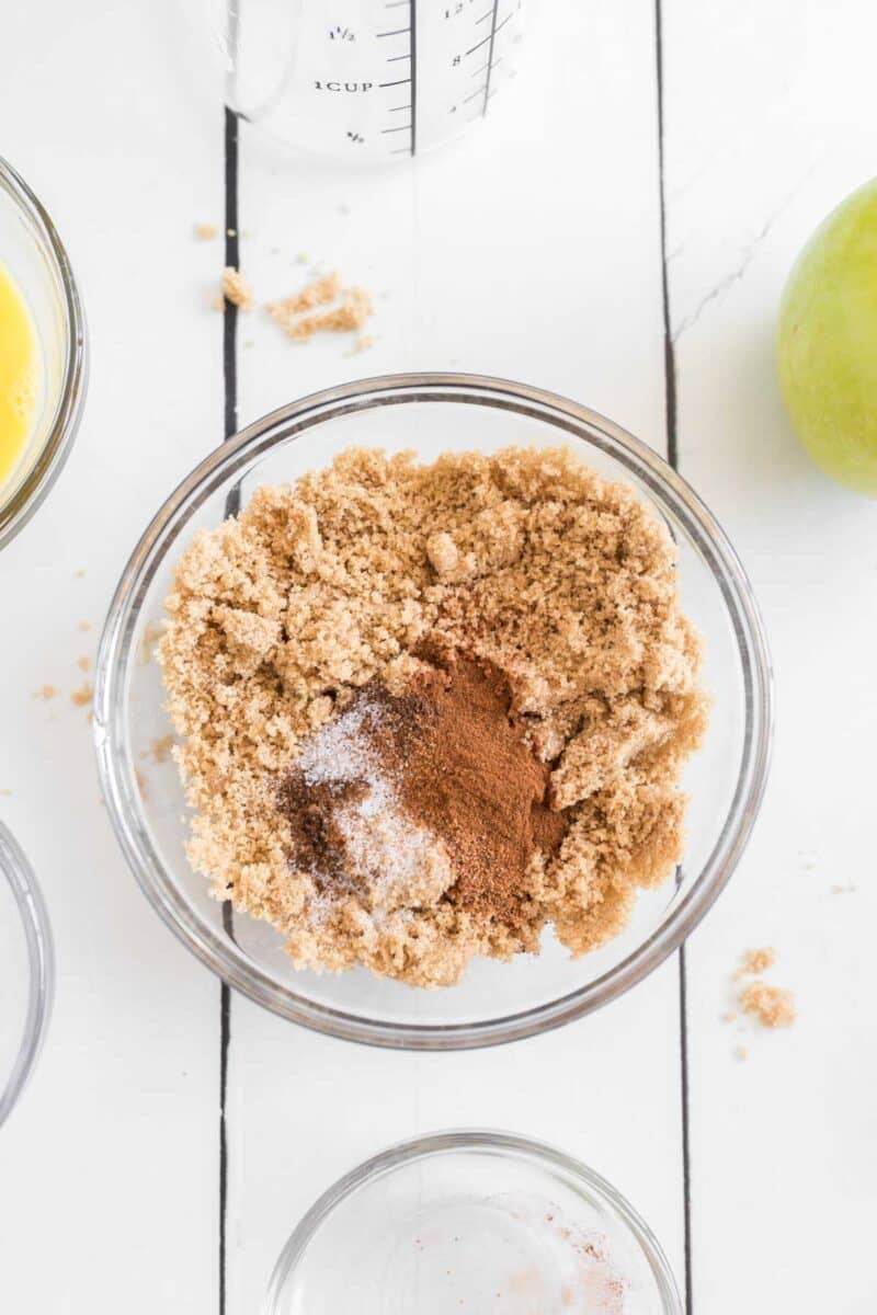 A glass bowl filled with brown sugar, cinnamon, and salt sits on a white surface, surrounded by other baking ingredients, including a green apple and a measuring cup.