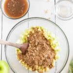 A glass bowl with chopped green apples and brown sugar being mixed with a spatula, surrounded by bowls of liquid ingredients on a white surface.