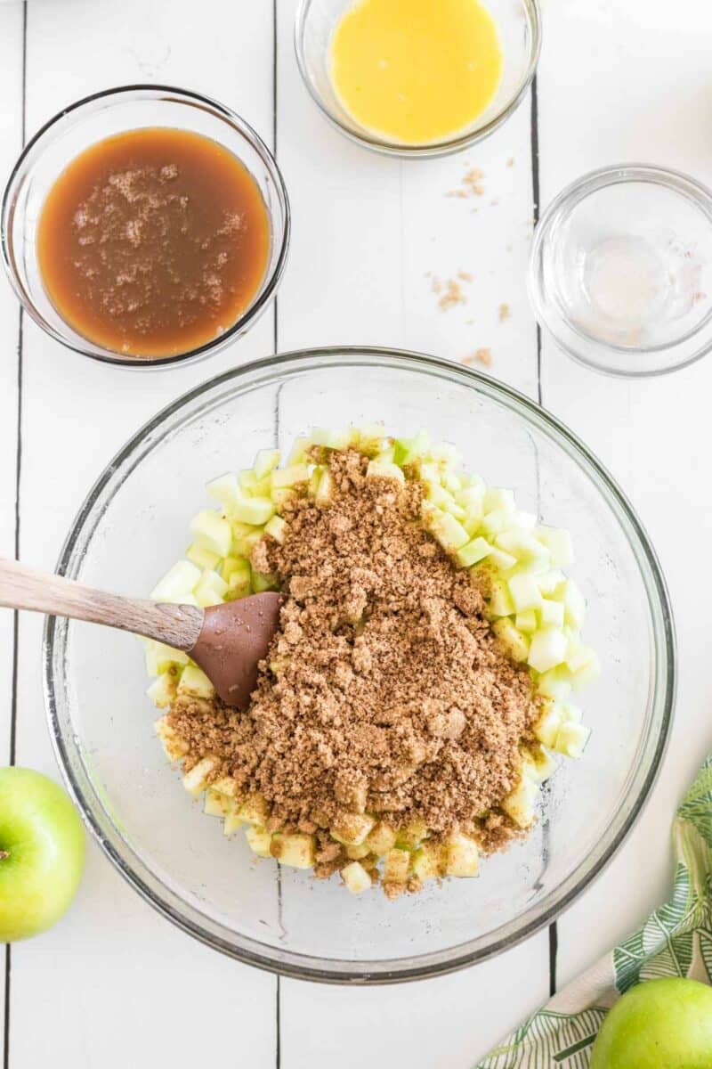 A glass bowl with chopped green apples and brown sugar being mixed with a spatula, surrounded by bowls of liquid ingredients on a white surface.
