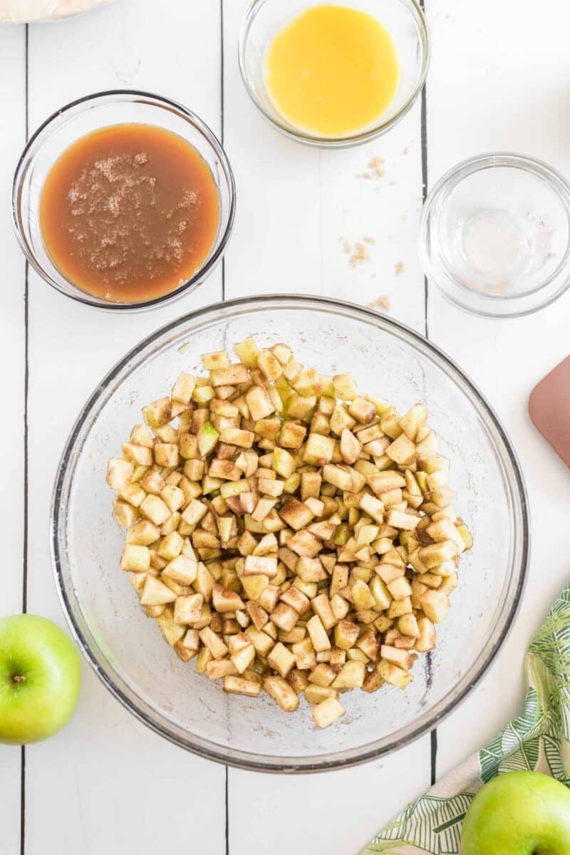 A glass bowl of diced apples coated with cinnamon sits on a white table, surrounded by bowls of brown liquid, yellow liquid, and a green apple.